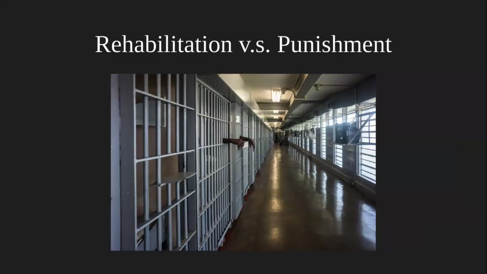 A still from the video presentation. It includes the words 'Rehabilitation v.s. Punishment' at the top of the image, and below there is a photo of a hallway of a prison facility. There are windows and technical equipment lining the right side of the hallway, and cells on the left. Two hands from a black individual rest through one of the cells.